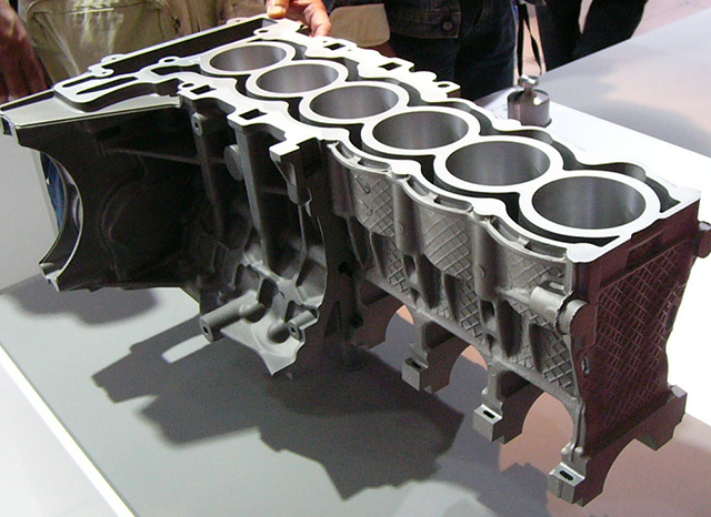 Un gros plan d"un bloc moteur en coupe, mettant en valeur la structure interne complexe et les al&eacute;sages des cylindres. Le composant m&eacute;tallique est expos&eacute; sur une table, plusieurs personnes l"observant en arri&egrave;re-plan. L"image met en &eacute;vidence les d&eacute;tails d"ing&eacute;nierie et de conception du moteur, gracieuset&eacute; d"une entreprise de moulage sous pression d"aluminium