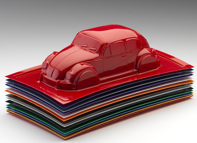 Un mod&egrave;le rouge brillant d&rsquo;une voiture classique, ressemblant &agrave; une Volkswagen Beetle, est plac&eacute; sur une pile soign&eacute;e de feuilles de plastique rectangulaires color&eacute;es. La voiture et les t&ocirc;les pr&eacute;sentent une surface brillante et r&eacute;fl&eacute;chissante, mettant en valeur le formage sous vide de plastique personnalis&eacute; avec une qualit&eacute; de classe mondiale sur un simple d&eacute;grad&eacute; allant du blanc au gris